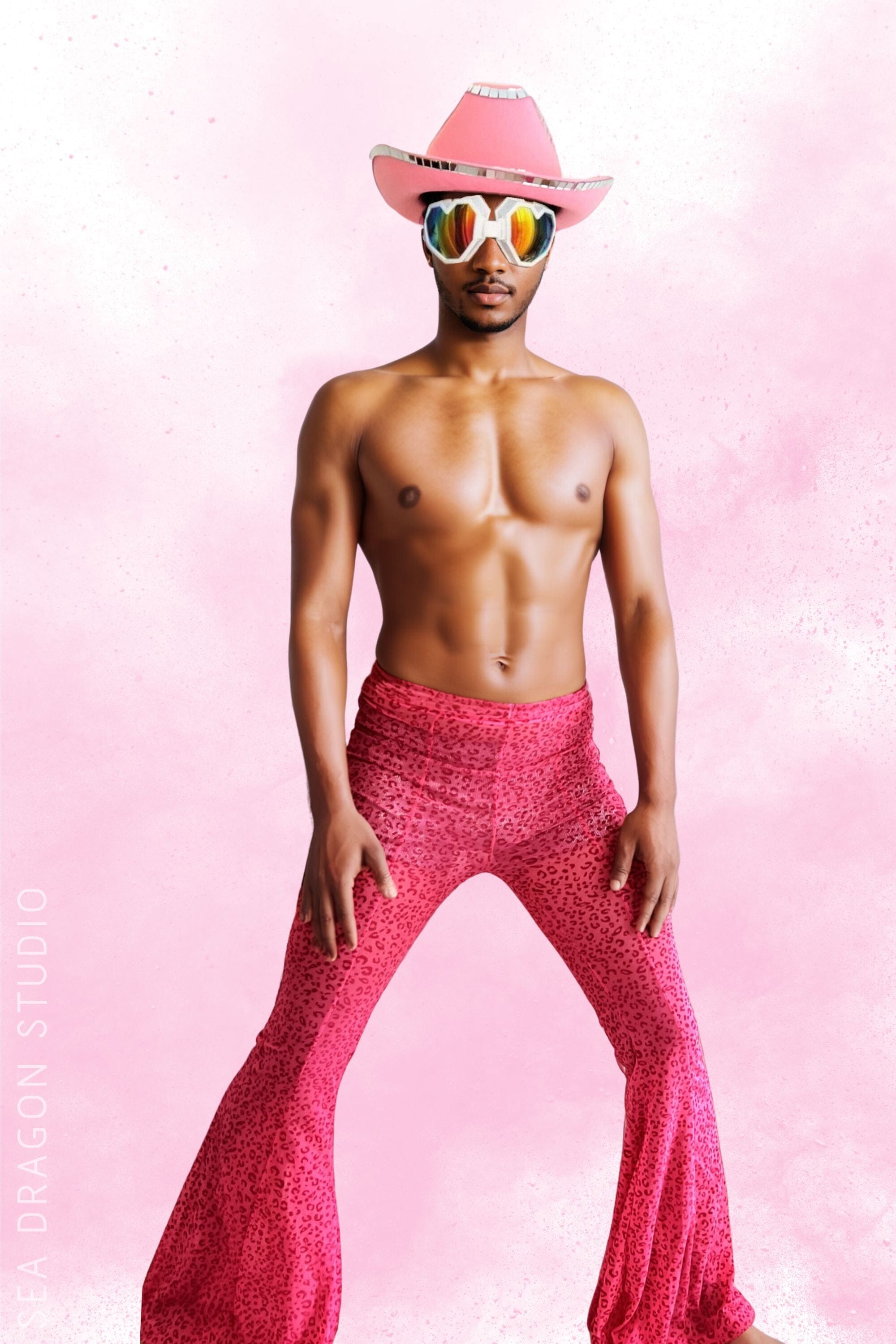 Model wearing men’s pink mesh flares perfect for Burning Man festival, showcasing high-waisted, flared pants with a vibrant pink leopard print, paired with a matching pink cowboy hat and rainbow sunglasses. Ideal for festival clothing mens, festival clothes for men, and festival wear for guys