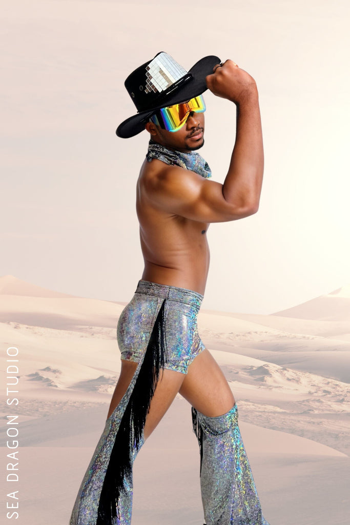 Man wearing holographic chaps and mens booty shorts with fringe, matching leg warmers, a mirrored cowboy hat, and reflective sunglasses. Perfect for Burning Man and festival fashion.
